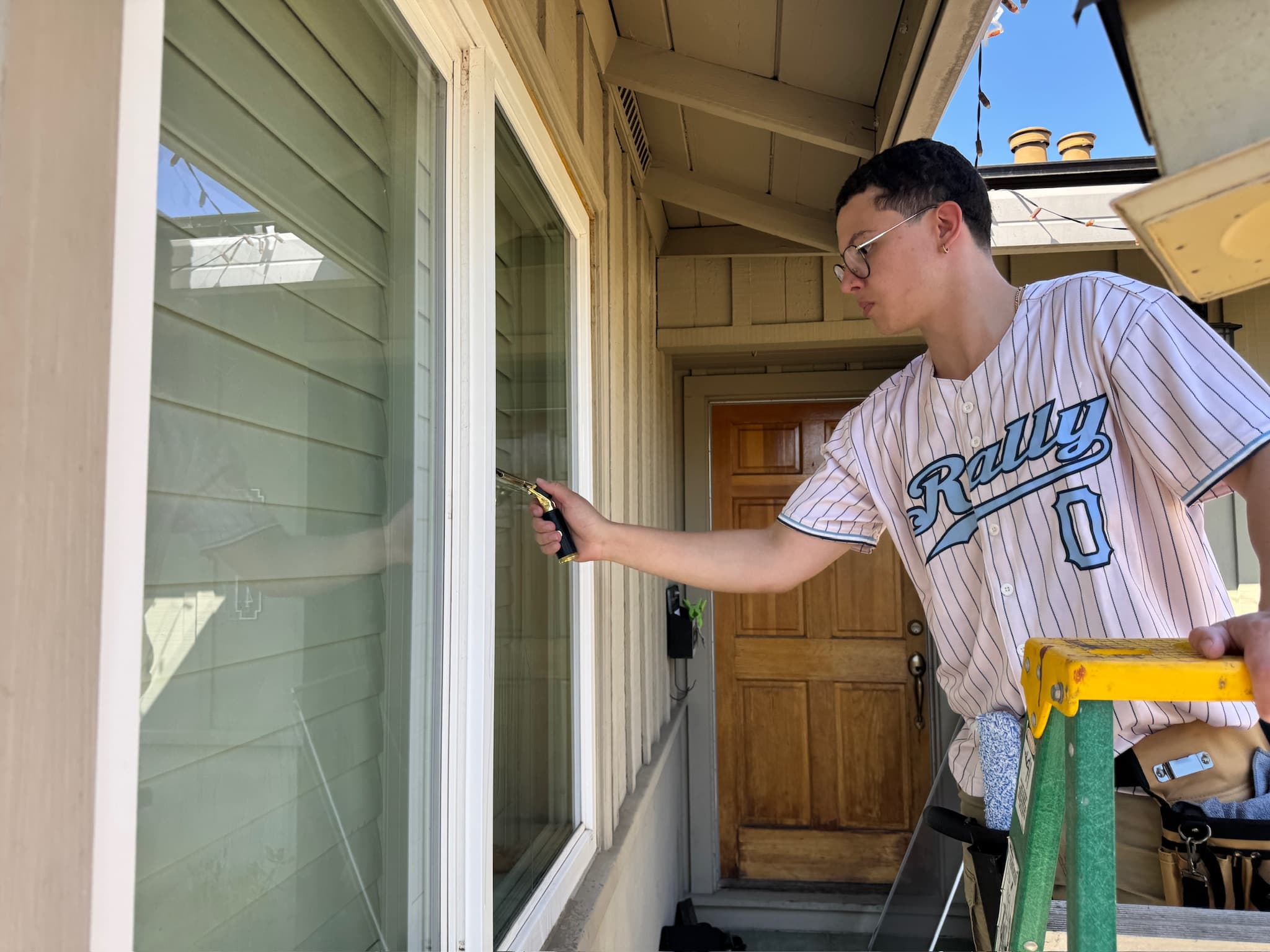 A Rally cleaner on a step ladder working a squeegee across the outside of a window.
