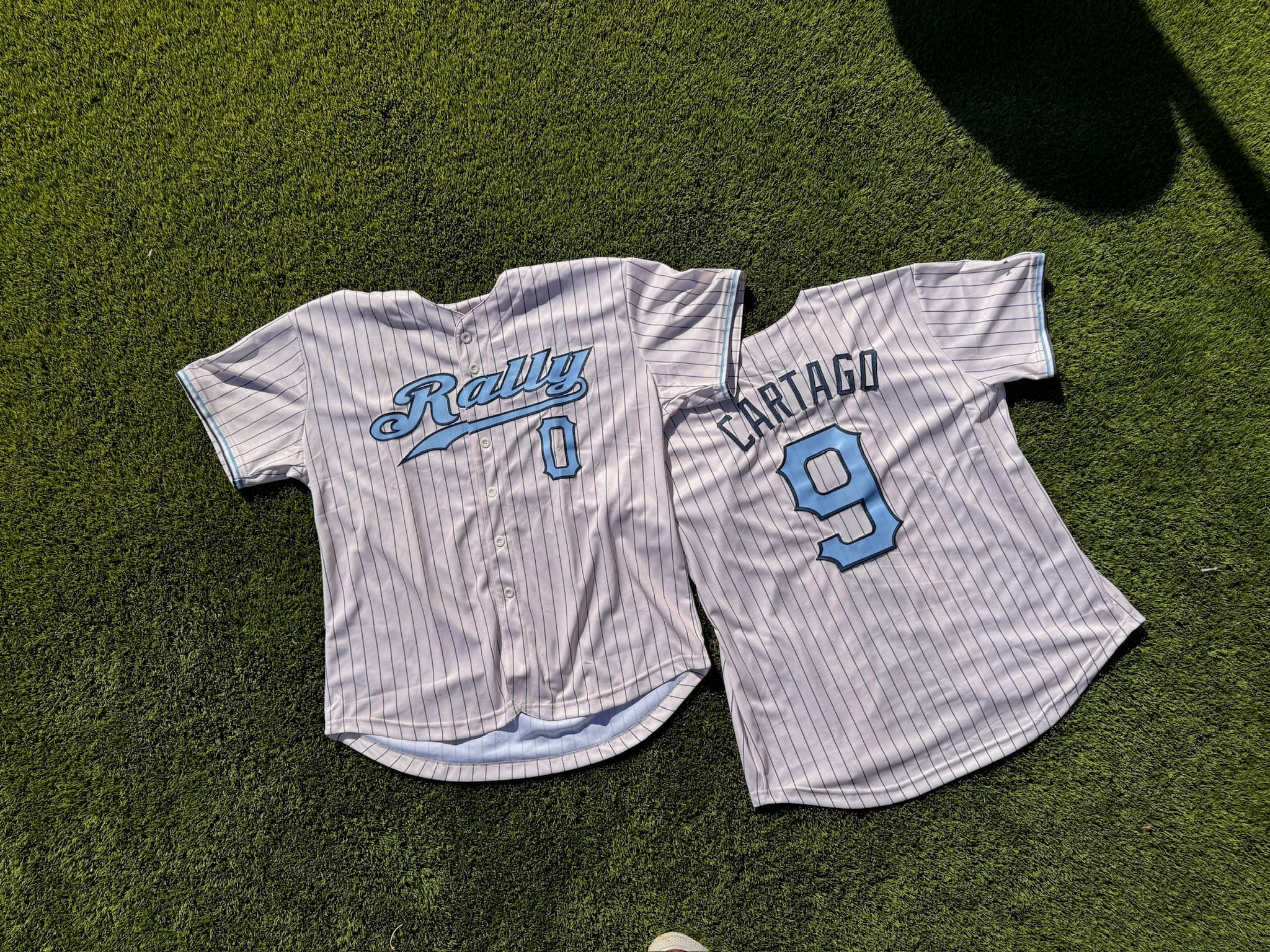 Two pinstriped Rally baseball jerseys laid out on grass — one front, showing the Rally script and the number 0, and one back, reading Cartago above the number 9.