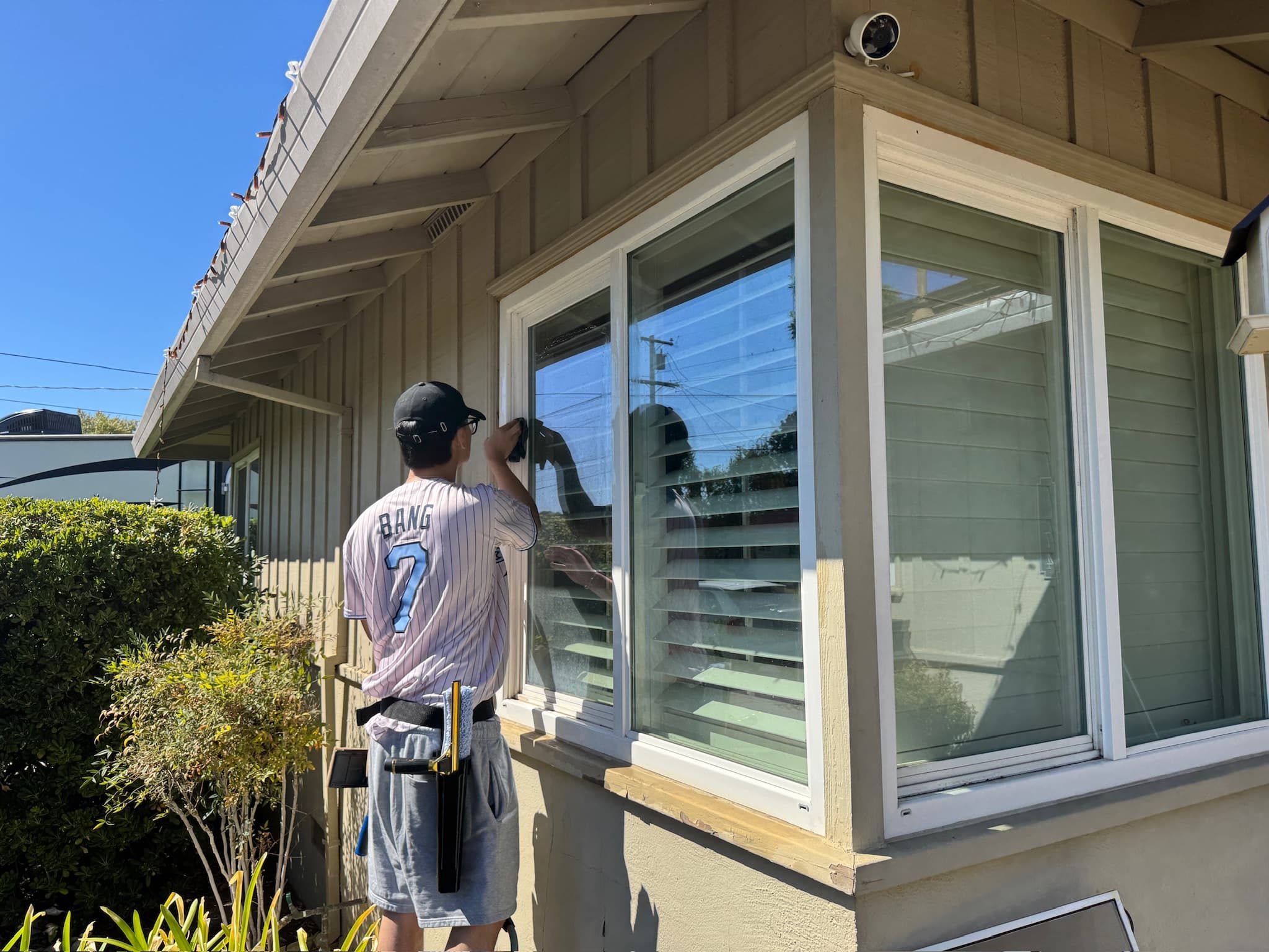 A Rally cleaner in a jersey and tool belt washing a large pane of exterior glass at a home.