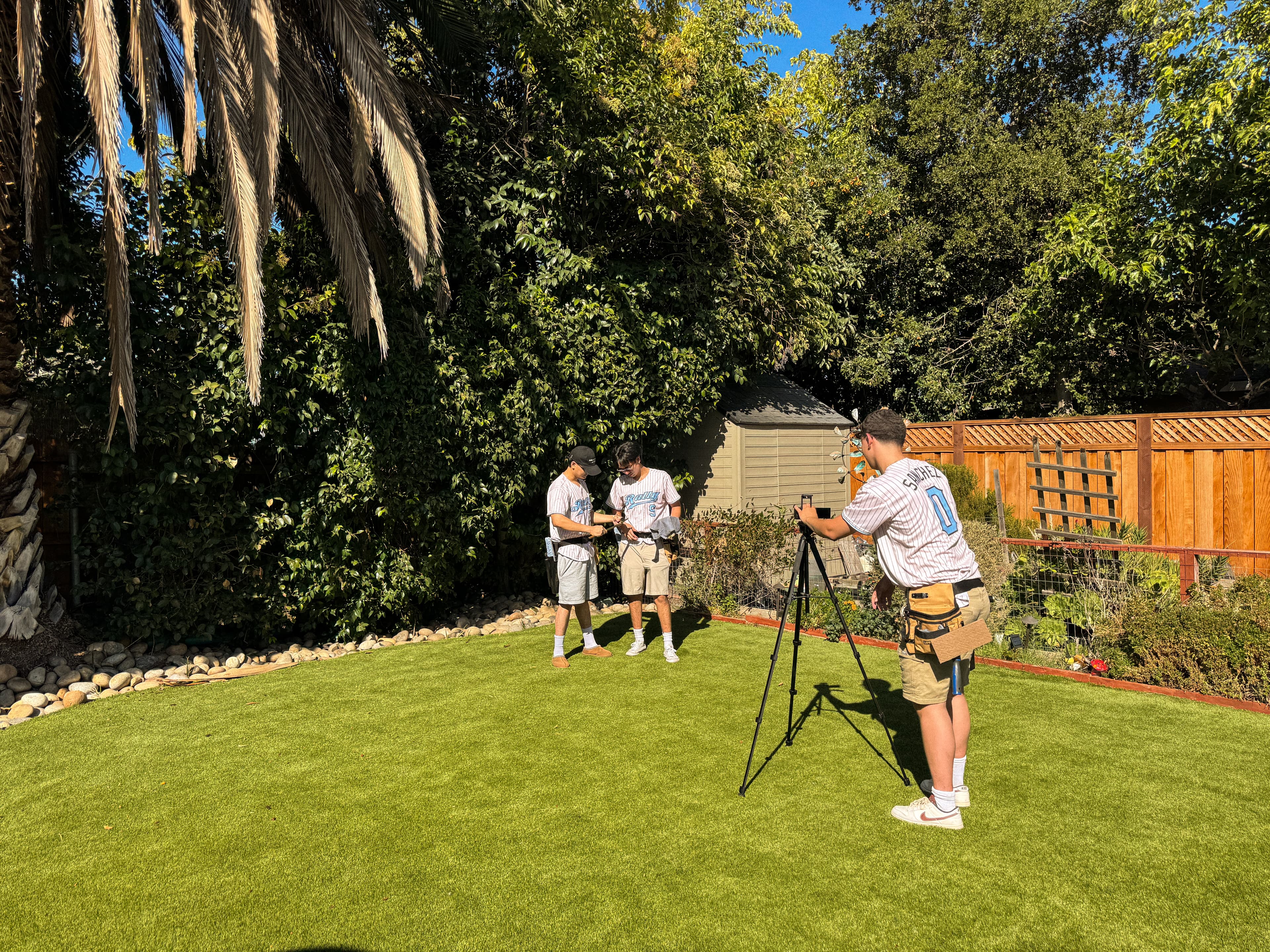 Three of the Rally crew in jerseys filming in a backyard — one lining up a phone on a tripod while the other two stand in frame.